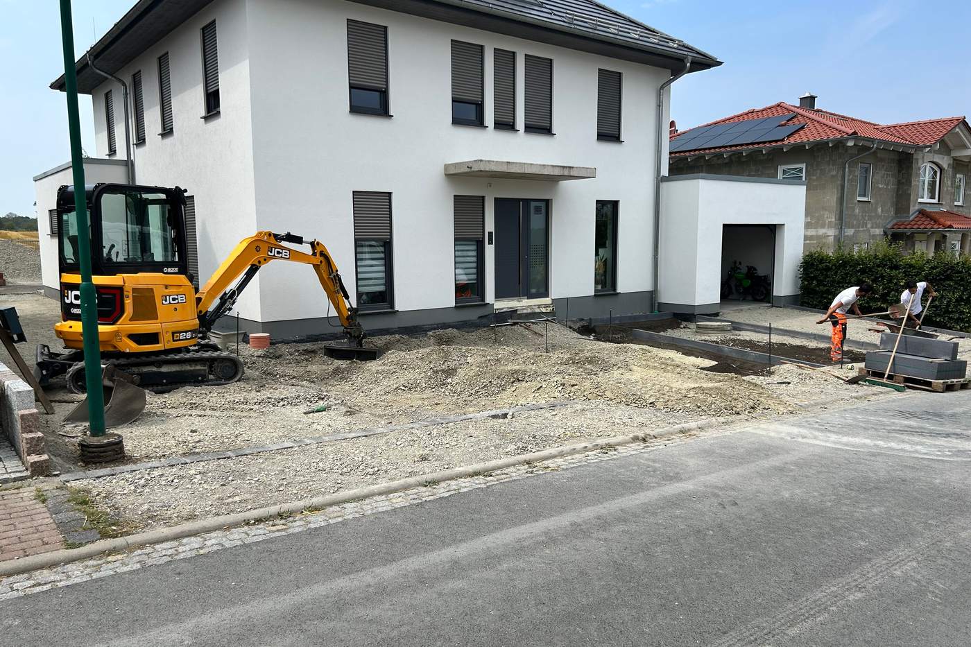 Kompaktbagger beim Aushub der Zufahrt an einem Neubau, daneben gestapelte Pflastersteine