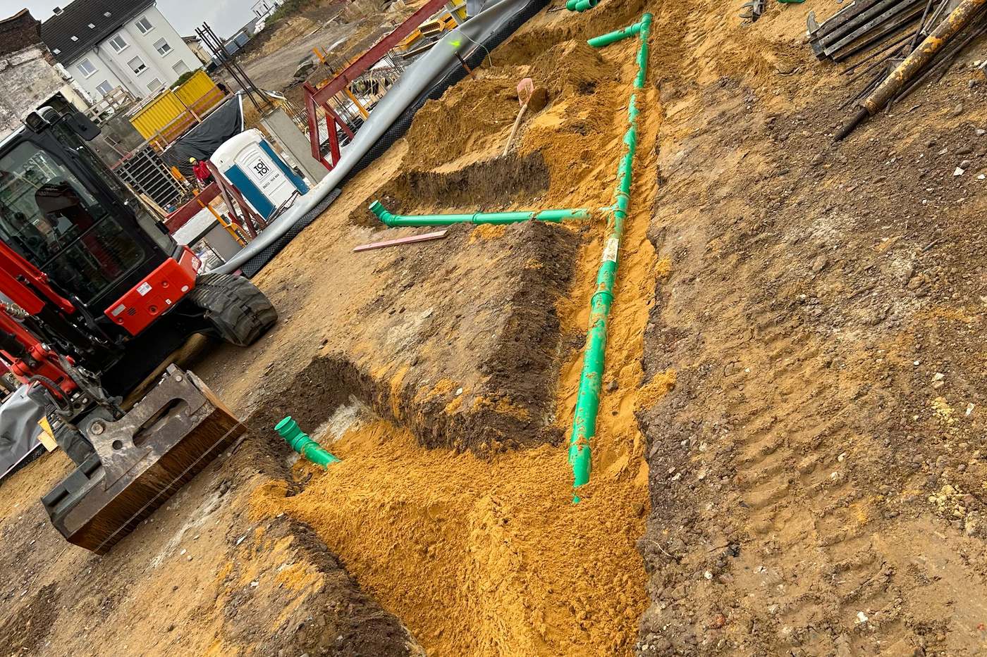 Leitungsgraben mit verlegten Kanalrohren, daneben ein Bagger auf der Baustelle