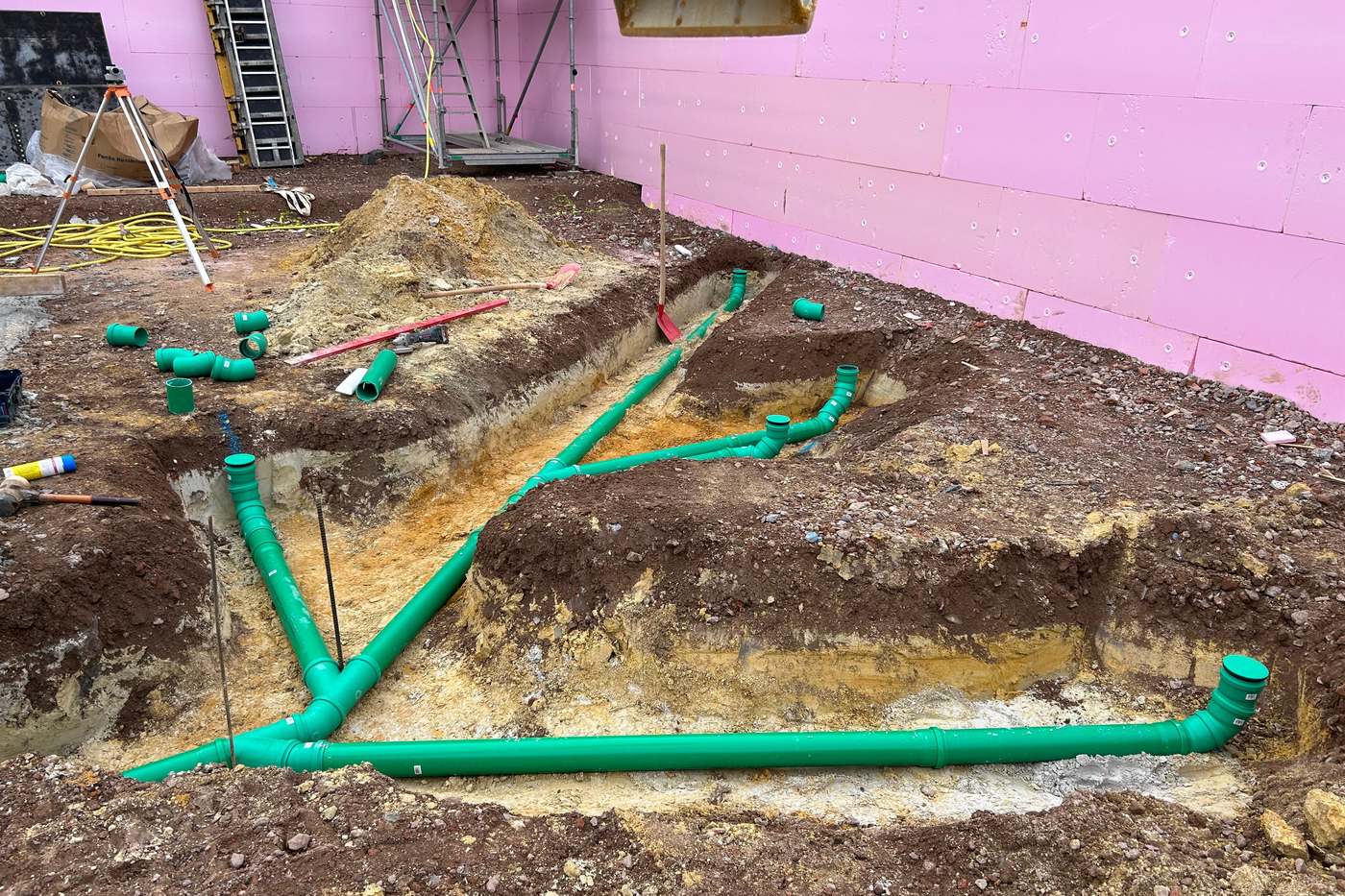 Verzweigte Grundleitungen aus grünem Kanalrohr im offenen Graben an einem Rohbau